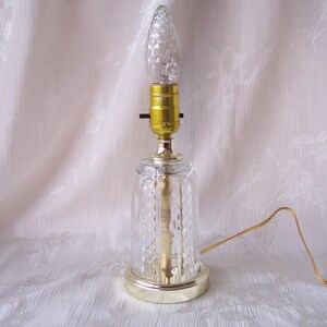 May include: A vintage table lamp with a clear, diamond-patterned glass base and a gold-colored metal base and accents. The lamp features a decorative, pointed lightbulb and a gold-colored cord.