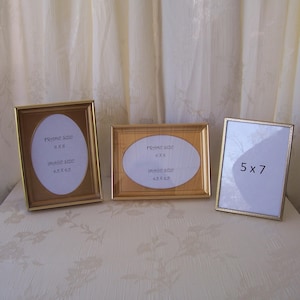 May include: Three gold-toned picture frames of varying sizes. Two frames have oval openings with text indicating frame and image sizes. The third frame is rectangular with a 5x7 opening. All frames are displayed on a patterned surface.