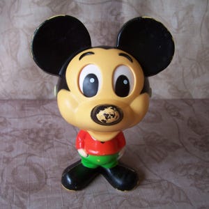 May include: A vintage plastic Mickey Mouse toy with black ears, yellow face, red shirt, and green pants. The toy has large eyes and a black nose.
