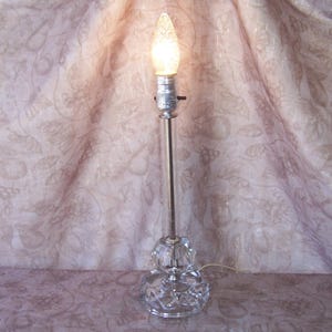 May include: A clear glass table lamp with a silver metal stem and base. The base has a cut-glass design. The lamp has a lit, teardrop-shaped bulb. The lamp is plugged in and ready to use. The lamp is a decorative lighting fixture.