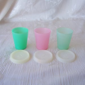 May include: Three small plastic cups in bright green, soft pink, and pale mint green, each accompanied by a matching circular white lid. The cups have a simple, tapered cylindrical shape with a smooth finish, arranged in a row with lids placed below.