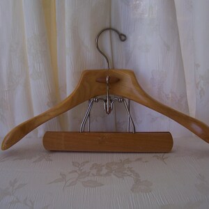May include: A wooden clothes hanger with a curved design and a metal hook. The hanger has a bar at the bottom for pants or skirts. The wood has a light brown color.
