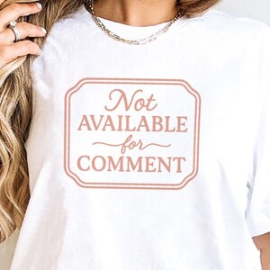 May include: White t-shirt featuring a light brown rectangular graphic with the words "Not AVAILABLE for COMMENT" in a decorative font. The text is centered. The shirt has short sleeves.