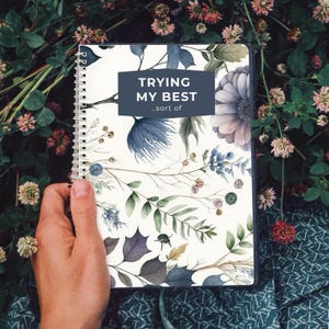 Peut inclure: Un carnet à spirales avec un motif floral. La couverture présente des illustrations à l'aquarelle bleues, violettes et vertes. Le texte "TRYING MY BEST .sort of" est dans un rectangle bleu foncé. Le carnet est tenu sur un fond de feuillage vert.