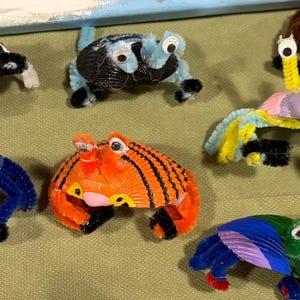 May include: A selection of handmade crab figurines in various colours. The crabs are made from pipe cleaners and other craft materials. Each crab has googly eyes and unique colour combinations, such as orange with black stripes or blue and yellow.