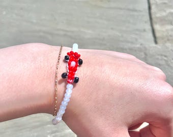 May include: A beaded bracelet featuring a red and black car design. The bracelet has a white and clear bead strand and a gold chain bracelet next to it. The car design is the focal point of the bracelet.