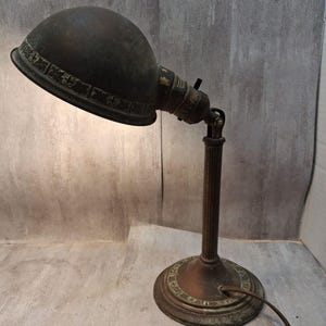 May include: A dark bronze-toned metal desk lamp featuring a dome-shaped shade with an embossed decorative border. The lamp has a fluted vertical stand and a circular base with matching embossed detailing. A power cord extends from the base.
