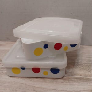 May include: Two white, square food storage containers with clear lids are stacked. The containers are decorated with colourful polka dots in yellow, red, and blue. The lids are slightly translucent.