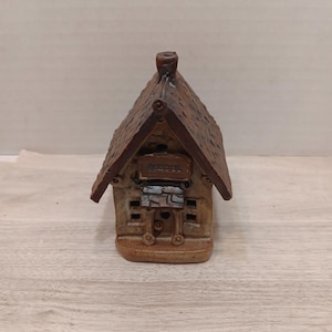 Puede incluir: Pequeña figura artesanal de cerámica que representa una escuela. El tejado marrón está texturizado, con una chimenea. La fachada muestra la palabra "SCHOOL". El edificio tiene un aspecto rústico y envejecido, con una base marrón claro y detalles de piedra.