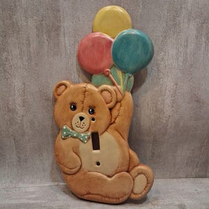 May include: A light brown teddy bear light switch cover featuring a green polka dot bow tie and three balloons in yellow, pink, and teal. The bear is depicted in a seated position with one arm raised to hold the balloons, and a vertical cutout.