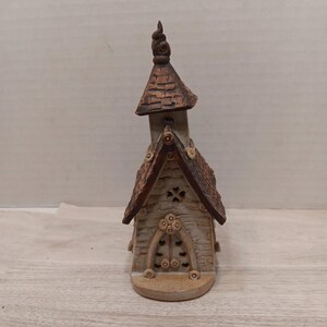 May include: A ceramic church figurine with a brown roof and steeple. The building has a light tan exterior with decorative details, including a cross-shaped window and arched doorway. The figurine is approximately 5 inches tall.