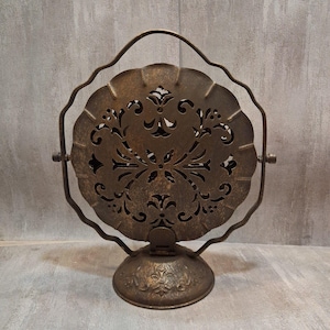May include: A circular, bronze-toned metal decorative object featuring intricate cut-out scrollwork patterns. It is suspended within a wavy, thin metal frame that attaches to a sturdy, embossed circular base, allowing for rotation.