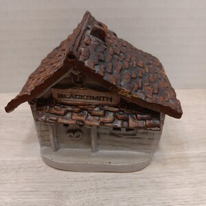 May include: A miniature ceramic blacksmith shop figurine. The roof is brown and textured, with a sign that reads "BLACKSMITH". The walls are light-colored with a textured appearance. The figurine has a rustic, handcrafted aesthetic.