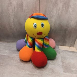 May include: A plush toy shaped like an octopus with a bright yellow head, a red nose, and embroidered blue eyes. It wears a small blue and orange cap. The eight tentacles feature alternating rainbow-striped patterns and solid colors including orange, red, green, purple, and pink.