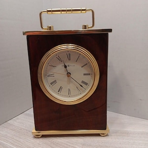 May include: A rectangular Howard Miller mantel clock with a dark brown wood finish, a gold-tone metal handle on top, and a gold-tone base. The clock face is white with black Roman numerals, black hands, and a gold-tone circular frame surrounding it.