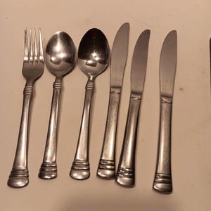 May include: A set of stainless steel silverware including a fork, two spoons, and three knives. The silverware has a simple, elegant design with a rounded handle and a decorative band near the base.