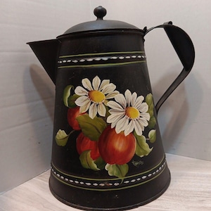May include: A black metal coffee pot featuring a hand-painted design of two white daisies with yellow centers and three red apples with green leaves. The pot has a lid with a small knob, a curved handle, and green and white decorative border stripes.