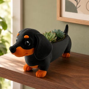 Peut inclure: Un pot de fleurs en forme de teckel noir et orange avec une finition mate lisse. La figurine de chien présente de grands yeux sombres, des oreilles tombantes et une petite ouverture sur le dos contenant deux petites plantes succulentes vertes.