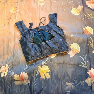 May include: A navy blue blouse with a floral pattern and a scalloped neckline. The blouse is set against a backdrop of a floral patterned fabric in shades of blue, peach, and yellow.