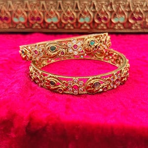 May include: Two gold bangles with intricate floral and geometric designs, adorned with red, green, and clear gemstones. The bangles rest on a vibrant pink surface, with a decorative gold box in the background. The jewelry is likely for special occasions.
