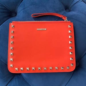 Puede incluir: Un bolso de mano naranja brillante con tachuelas cuadradas plateadas en los bordes. El bolso tiene un cierre de cremallera y una peque&ntilde;a correa de mu&ntilde;eca. El nombre de la marca, REBECCAMINKOFF, se muestra en una peque&ntilde;a barra plateada.