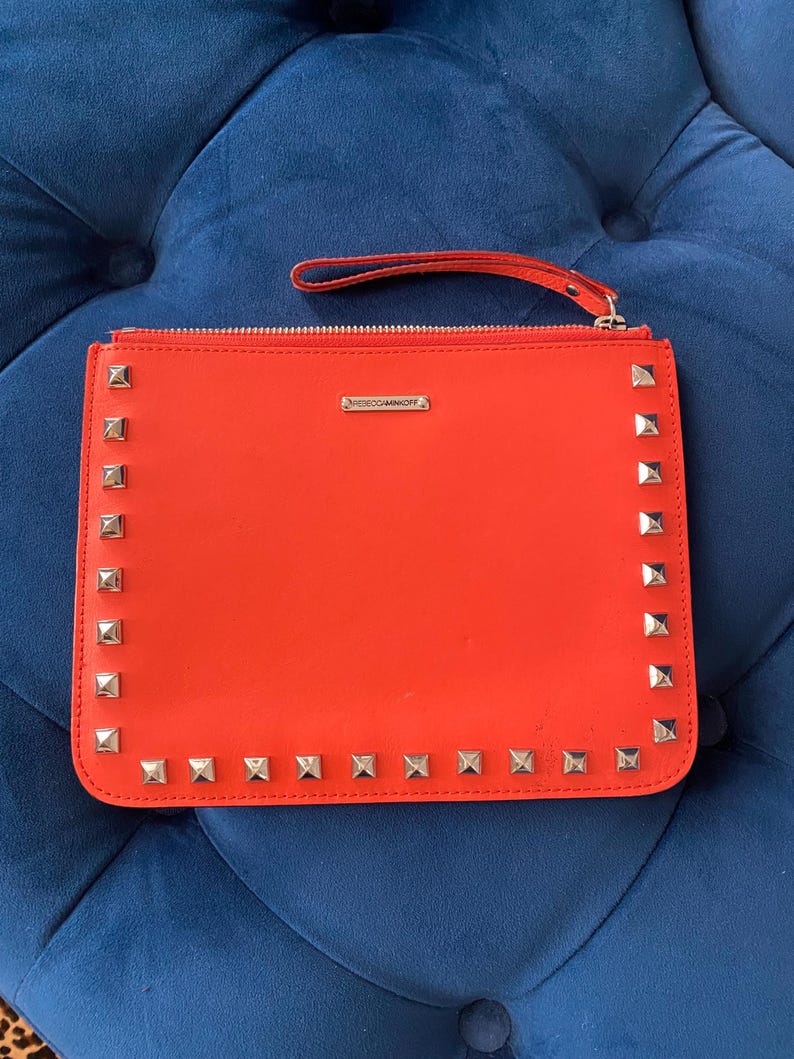 Puede incluir: Un bolso de mano naranja brillante con tachuelas cuadradas plateadas en los bordes. El bolso tiene un cierre de cremallera y una peque&ntilde;a correa de mu&ntilde;eca. El nombre de la marca, REBECCAMINKOFF, se muestra en una peque&ntilde;a barra plateada.