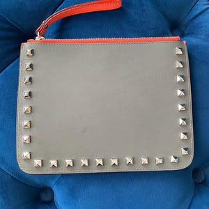 Puede incluir: Clutch gris con tachuelas cuadradas plateadas en los bordes. El clutch tiene una cremallera roja y una correa de mu&ntilde;eca roja a juego. El clutch es de forma rectangular.