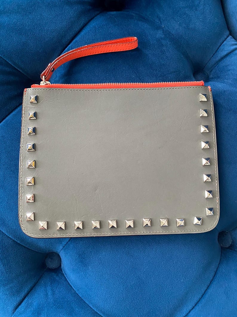 Puede incluir: Clutch gris con tachuelas cuadradas plateadas en los bordes. El clutch tiene una cremallera roja y una correa de mu&ntilde;eca roja a juego. El clutch es de forma rectangular.