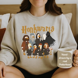 May include: A light gray sweatshirt with the word "Honkwarts" in orange lettering. The sweatshirt features a cartoon illustration of ducks dressed as characters from a popular fantasy series. A white mug with the text "Hand-drawn by a real Potterhead" is held in the hand.