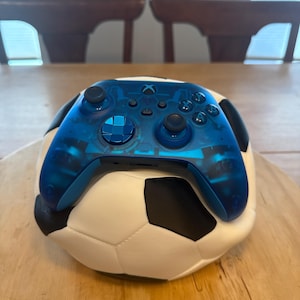 Puede incluir: Un mando Xbox azul transparente descansa sobre un balón de fútbol. El mando revela componentes internos. El balón es blanco con pentágonos negros. Sobre una superficie de madera.