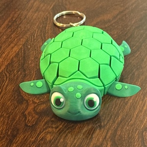 May include: A green turtle keychain with a gold-colored ring. The turtle has a geometric shell pattern and large, friendly eyes. The keychain is made of a solid material and is designed to be durable.