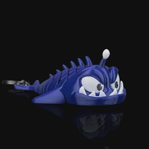 May include: A blue, cartoonish fish-like creature keychain with white teeth and eyes. The creature has a spiky dorsal fin and a small white bulb on its head. The keychain is made of a 3D-printed material and is set against a black background.