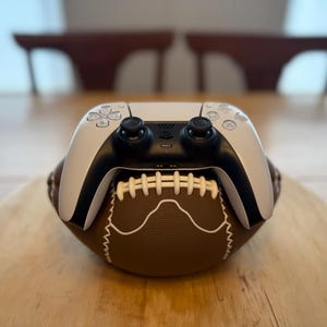 Puede incluir: Un mando blanco y negro de PlayStation 5 descansa sobre un balón de fútbol americano marrón. El balón tiene costuras blancas y está sobre una superficie de madera. El fondo está borroso.