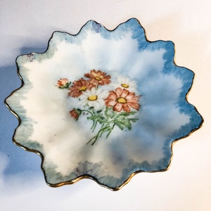 May include: A white porcelain dish with a scalloped edge and a gold trim. The dish features a hand-painted design of a bouquet of white and orange flowers.