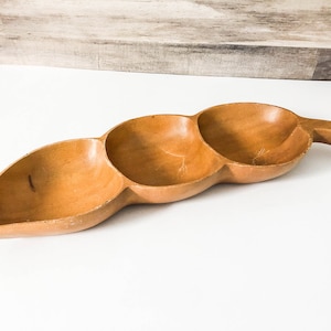 May include: A wooden serving dish with three separate compartments, shaped like a pea pod with a handle. The dish is made of light brown wood and has a natural finish.