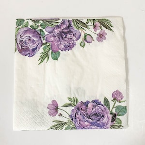 Puede incluir: Servilleta de papel blanca con un dise&ntilde;o floral morado. La servilleta presenta un borde de flores y hojas moradas, con una flor morada m&aacute;s grande en la esquina inferior derecha.