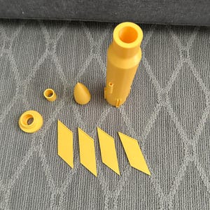 May include: A selection of yellow 3D-printed components, including a cylindrical part, a cone-shaped piece, and several diamond-shaped elements. The items are arranged on a grey patterned surface.
