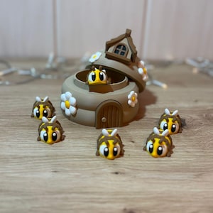 May include: A whimsical, tan-colored bee house with a small, open door and a rooftop. The house is decorated with white daisy accents. Several yellow and black bee figurines are scattered around the house, creating a playful scene.