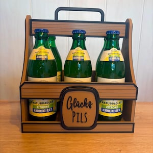 Könnte beinhalten: Ein Bierkasten aus Holz mit schwarzem Griff und Zierleisten, der sechs grüne Glasflaschen alkoholfreies Naturradler Zitrone Bier enthält. Der Kasten trägt die Aufschrift "Glücks Pils".