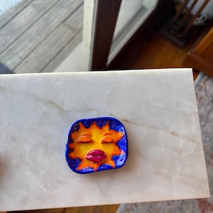 May include: A small, square ceramic dish with a sun face design. The sun has closed eyes, a large mouth, and is surrounded by yellow rays. The dish has a blue border and is resting on a white marble surface.