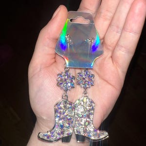 Puede incluir: Pendientes colgantes con diseño floral y colgantes de botas de vaquero. Los pendientes están adornados con pedrería iridiscente, creando un efecto brillante. Los pendientes están sujetos a una tarjeta holográfica.