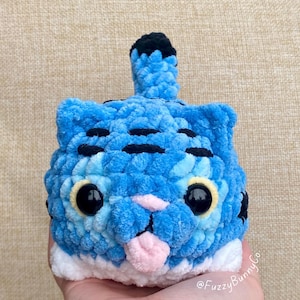 Puede incluir: Un peluche de ganchillo azul con forma de gato con rayas negras, grandes ojos negros, nariz rosa y lengua rosa hacia fuera. La base es blanca y el texto @FuzzyBunnyCo es visible en la esquina inferior derecha del peluche.