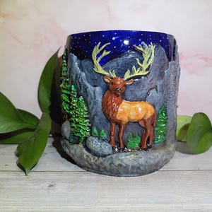 May include: A hand-painted ceramic mug featuring a detailed elk standing in a mountain scene. The mug has a dark blue night sky with stars, gray mountains, green trees, and a brown and tan elk. The mug is approximately 4 inches tall.