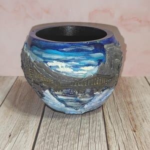 May include: A round, hand-painted ceramic pot with a detailed landscape design. The pot features a bridge over water, with blue and white painted sky and water, and gray mountains. The interior is black.