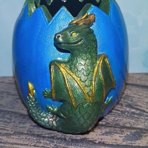 May include: A hand-painted ceramic dragon emerging from a blue egg. The dragon is green with gold accents on its wings and horns. The egg has a jagged top edge with green trim.