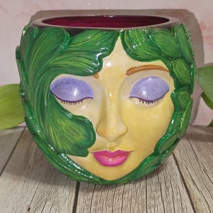 May include: A ceramic planter with a face design. The face has closed purple eyes, a yellow complexion, and pink lips. Green leaves frame the face, and the interior rim is a deep burgundy color. The planter sits on a wooden surface.