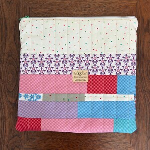 Puede incluir: Una bolsa con cremallera de patchwork con una variedad de telas coloridas. La sección superior presenta un fondo crema con lunares rojos, verdes y rosas. Debajo, hay un estampado floral, y la sección inferior es una mezcla de bloques rosas, rojos, azules y morados.