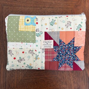 Puede incluir: Un estuche acolchado con cremallera y diseño de patchwork. El estuche presenta cuadrados de tela verde, amarilla y crema con estampados de conejos y flores. Un gran diseño de estrella está en el lado derecho, y una pequeña etiqueta está cosida en la parte delantera.