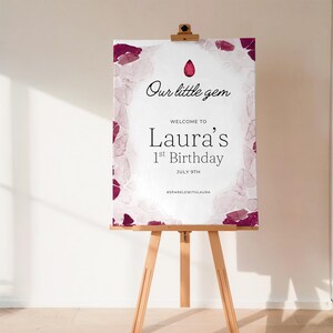 May include: A rectangular birthday sign on a wooden easel featuring a white background with dark pink watercolor accents along the edges. The text reads Our little gem, WELCOME TO Laura's 1st Birthday, JULY 9TH, and #SPARKLEWITHLAURA.