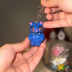 May include: A blue cat-shaped keychain with a silver clasp. The cat has large orange eyes, pink ears, and a small pink nose. The keychain is held in front of a blurred background.